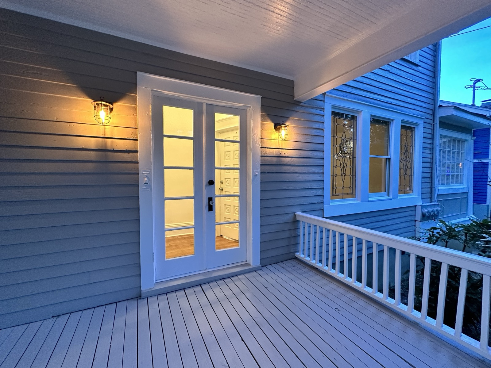 Front porch at dusk with white French doors, lit lantern sconces on gray siding, and leaded-glass windows beyond