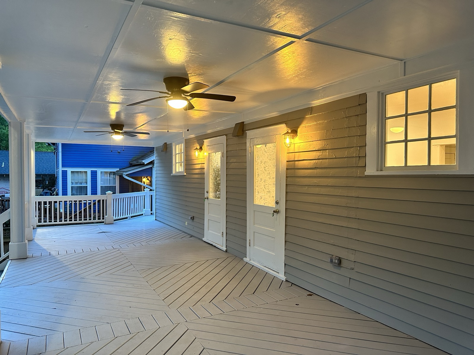 Covered upper rear porch at dusk with two ceiling fans, herringbone-laid decking, and doors lit by sconces